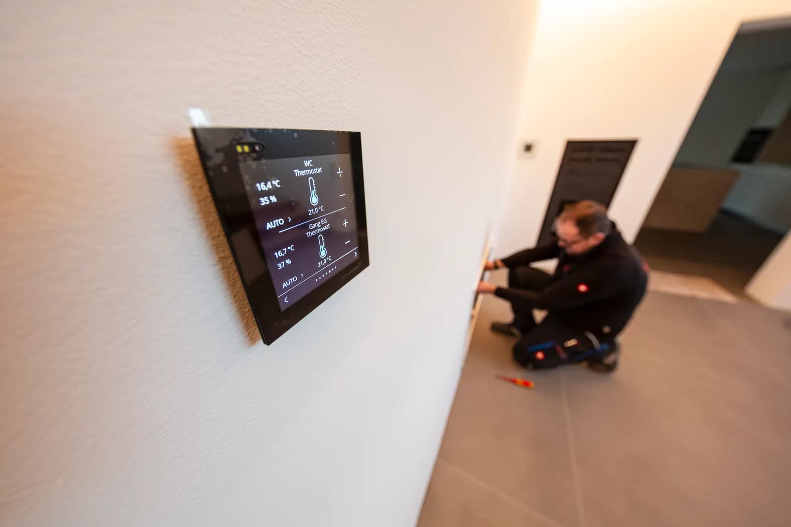 Wandmontiertes Smart-Home-Bedienpanel zur Steuerung von Heizung und Licht, im Hintergrund ein Monteur bei der Installation